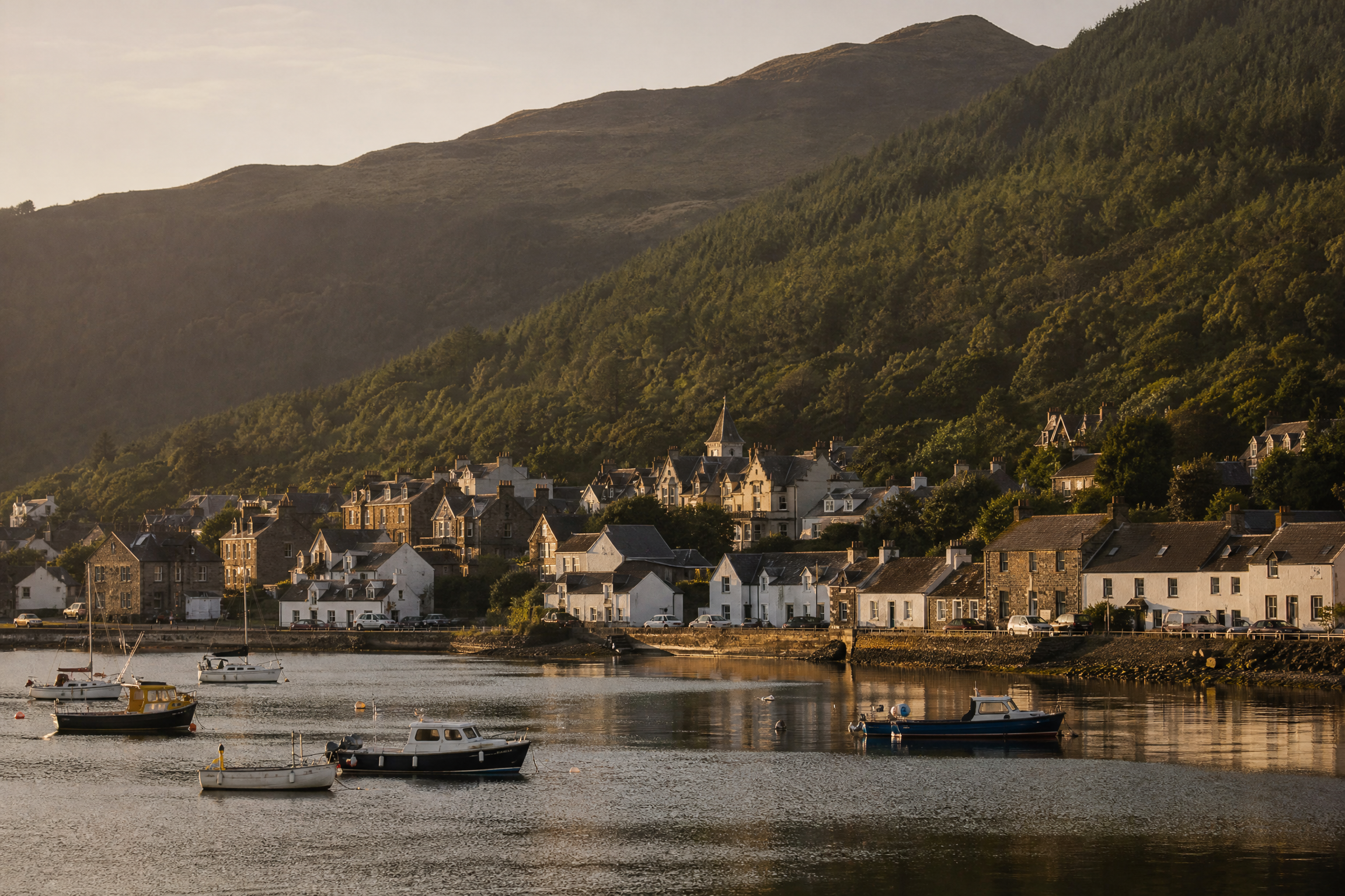 Brodick village waterfront at golden hour, small boats moored in the bay, forested hills behind, Isle of Arran, Scotland
