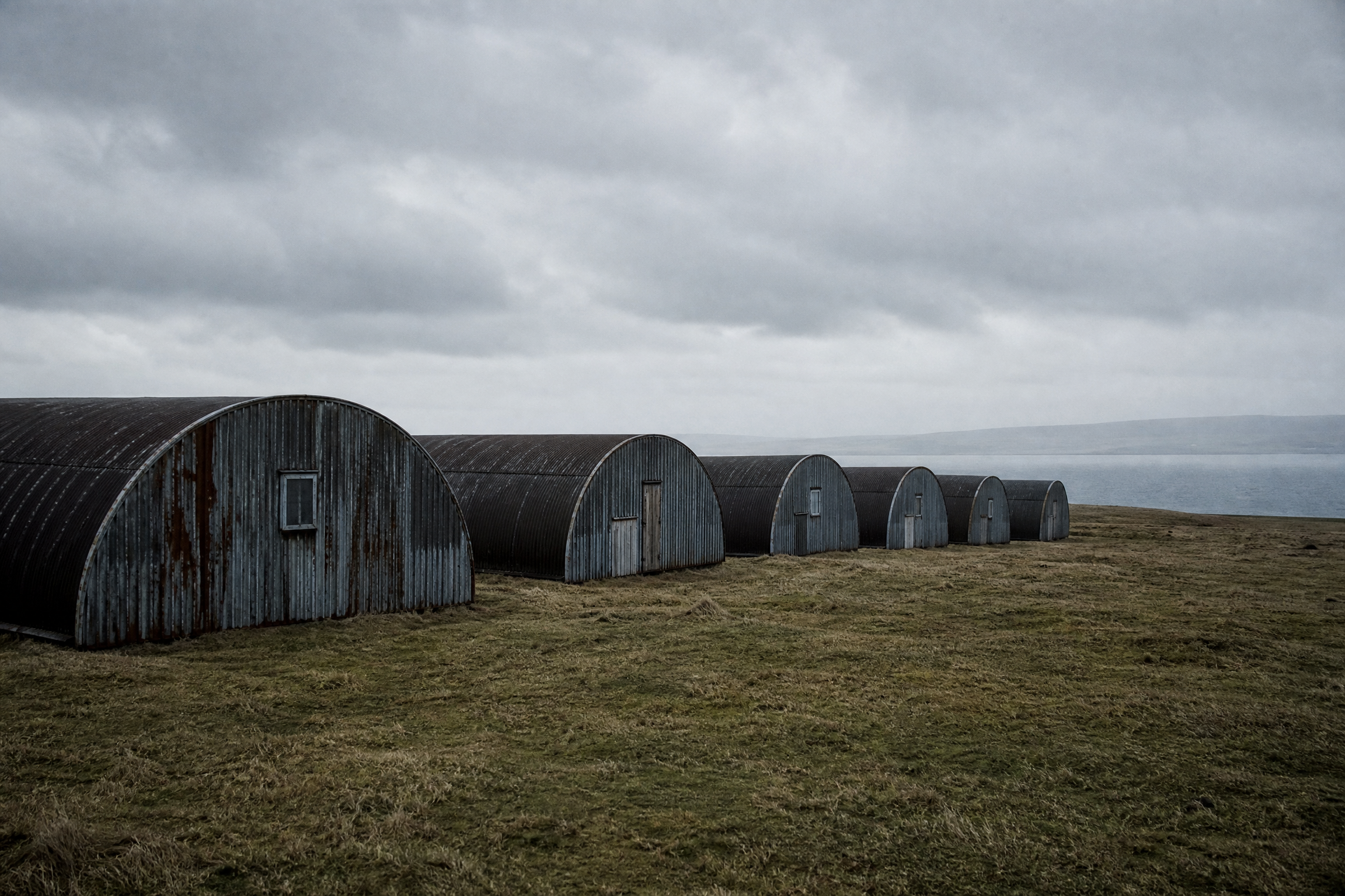 Row of weathered corrugated Nissen huts on a windswept Orkney clifftop, grey sea behind, evoking the wartime Camp 60 site on Lamb Holm
