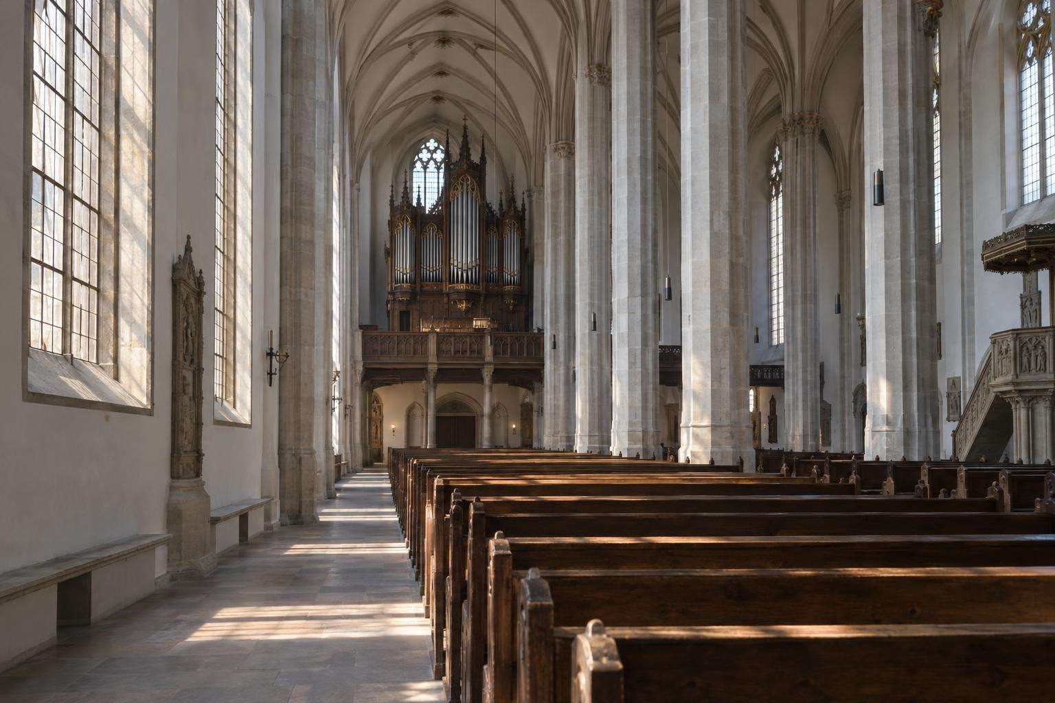 The nave of a Gothic Lutheran hall church in Leipzig in early-morning light, with wooden pews leading towards the organ gallery.