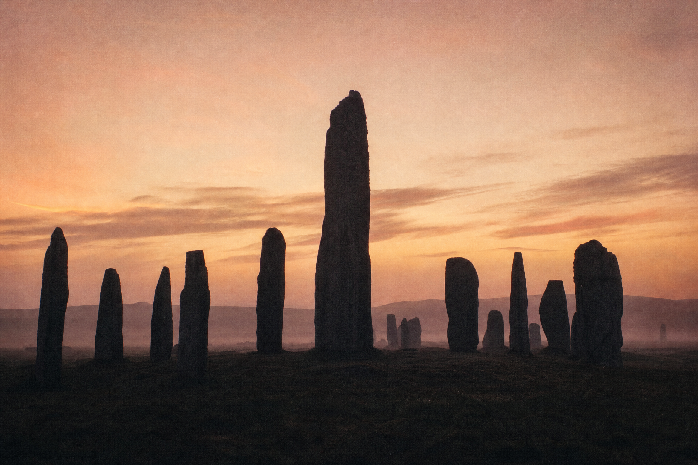 Dawn silhouette of the full Callanish stone circle and tall central monolith against a rose and amber pre-dawn sky with moorland mist