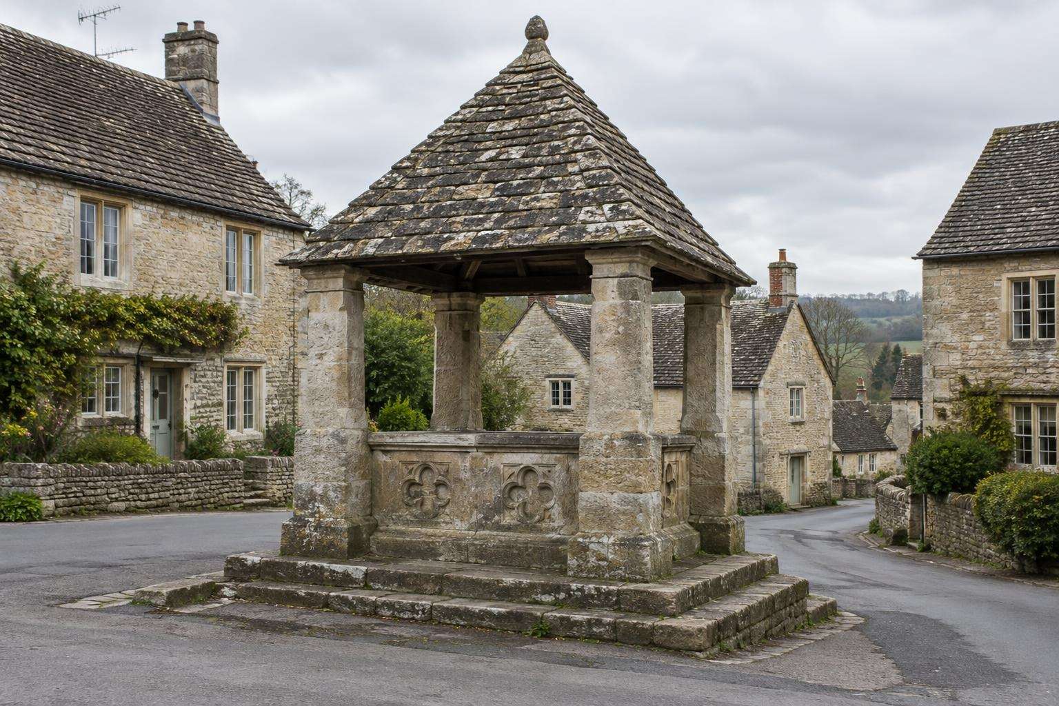 A late-medieval market cross with a stone-tiled pyramid roof on four piers in a Wiltshire village market place.