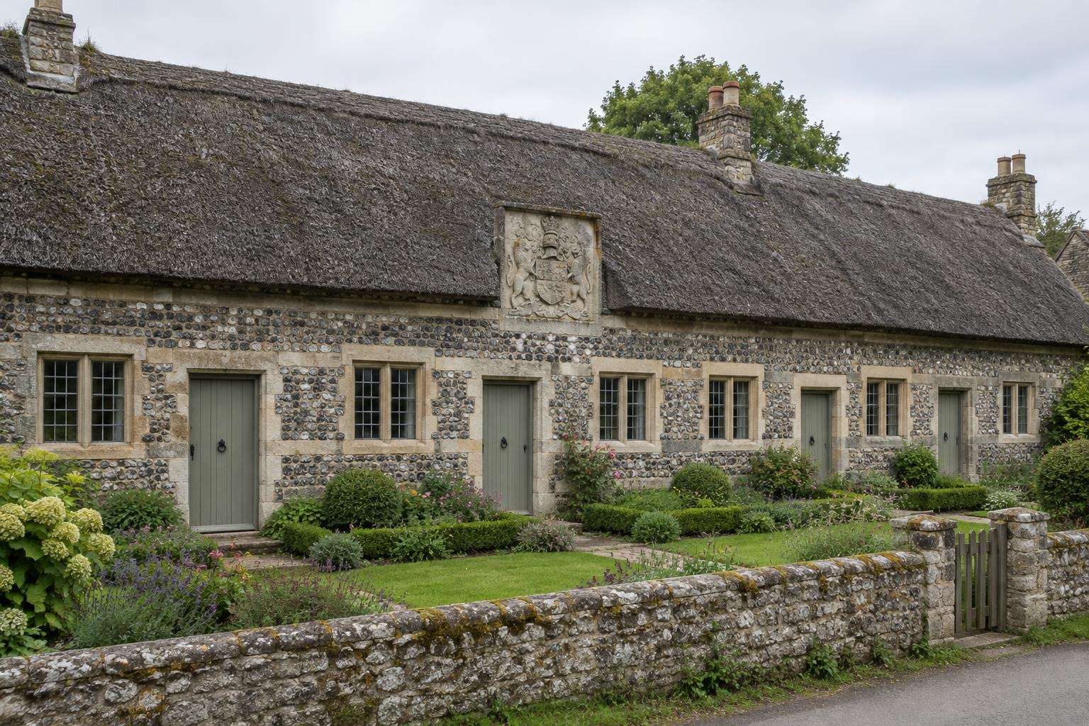 A low row of thatched flint-and-stone almshouses with a carved heraldic panel in a Dorset estate village.
