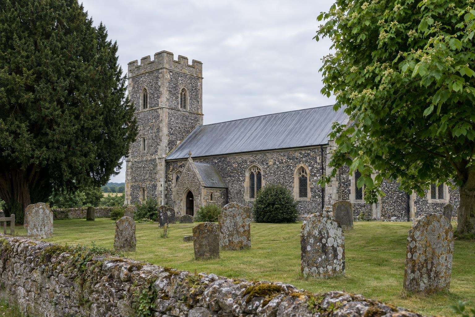 A medieval flint and stone parish church with a battlemented tower and lichen-covered headstones in a Wiltshire churchyard.