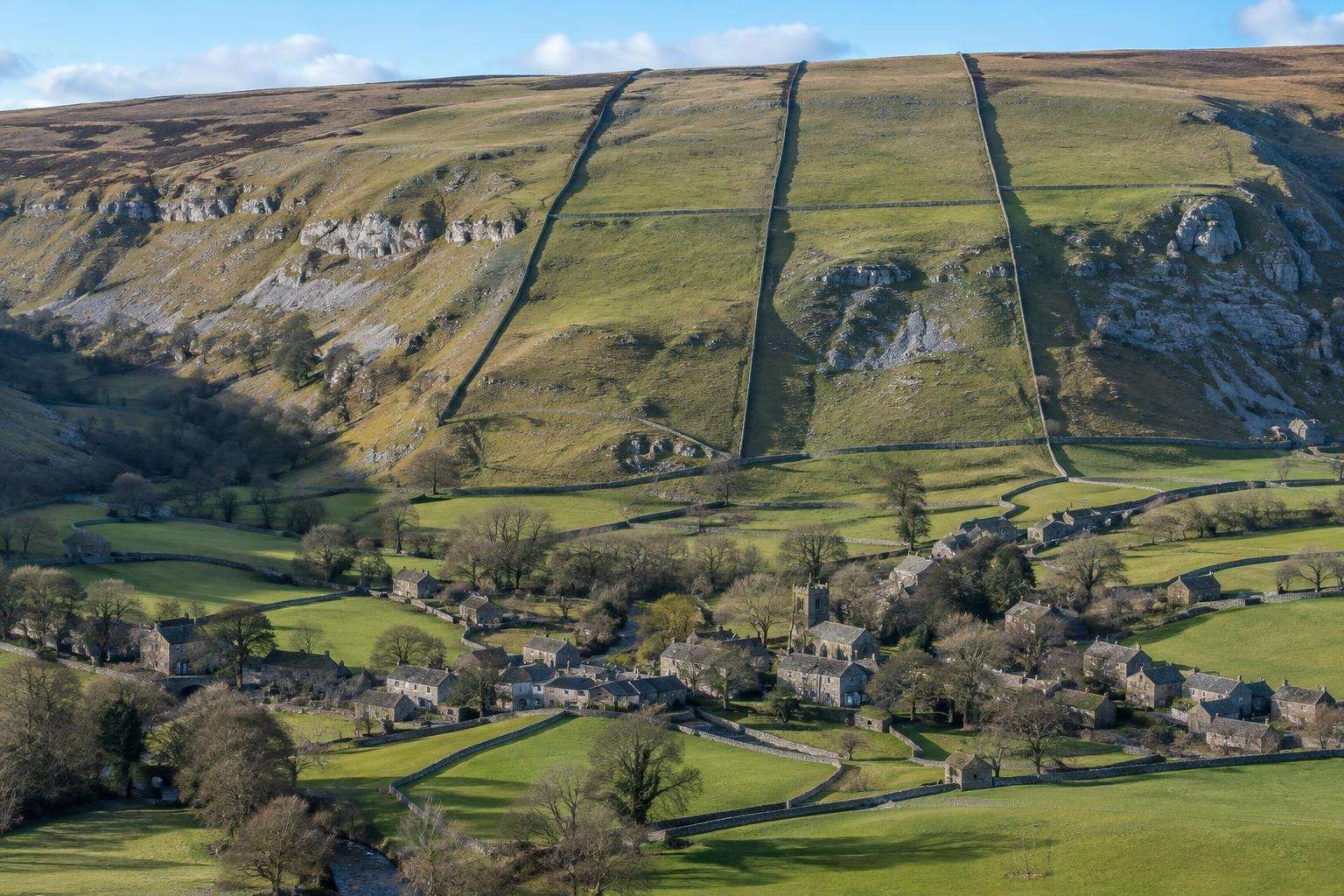 Straight enclosure walls running up a fellside above small irregular fields around a stone village in Wharfedale, Yorkshire Dales.