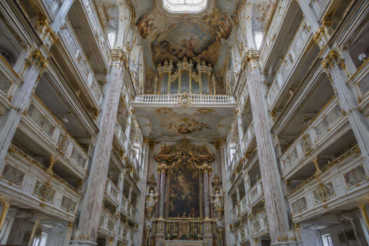 A tall, narrow Baroque palace chapel with white and gold galleries rising in tiers and an organ set high under the ceiling, lit from above