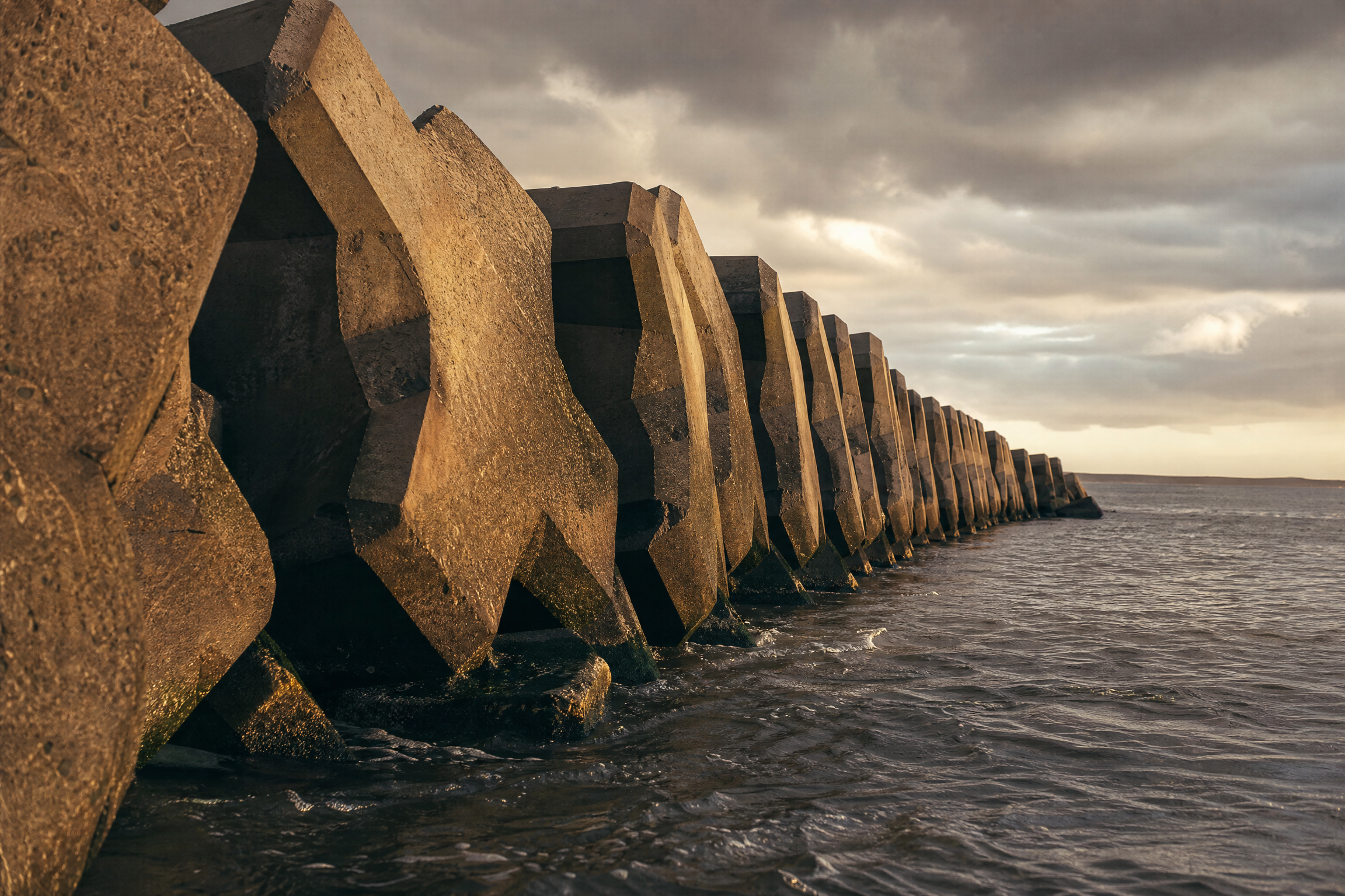 Row of massive concrete blocks forming a Churchill Barrier, tidal water at the base, under a moody sky