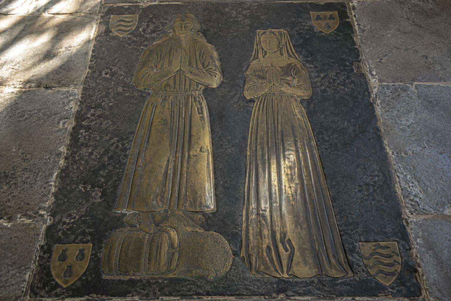 A worn medieval brass of a wool merchant and his wife, his feet on a woolpack, set in a Cotswold church floor.